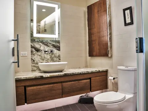 A bathroom with a floating walnut vanity, a vessel sink on a stone counter and a lit mirror