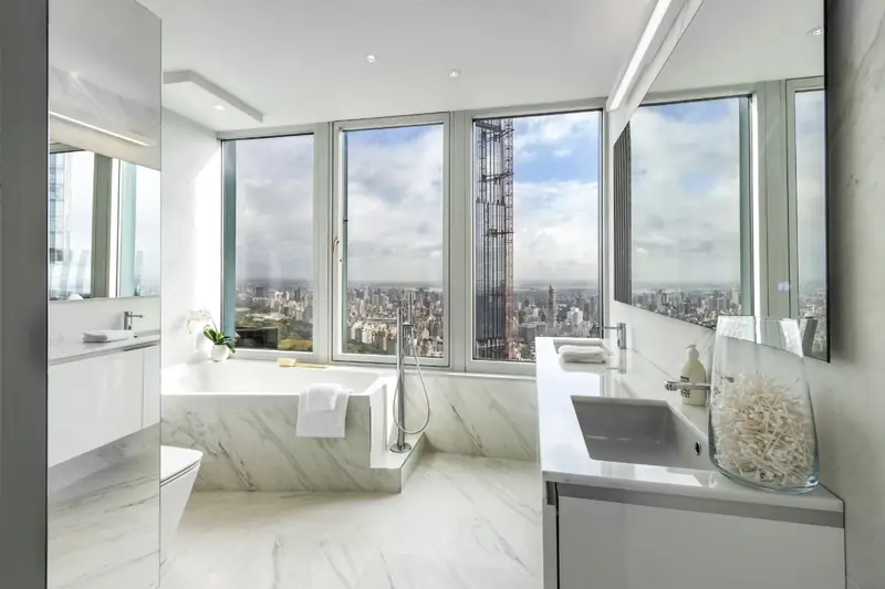 A white marble bathroom with a tub in a marble deck under a wall of windows onto a park and skyline, and a long white vanity