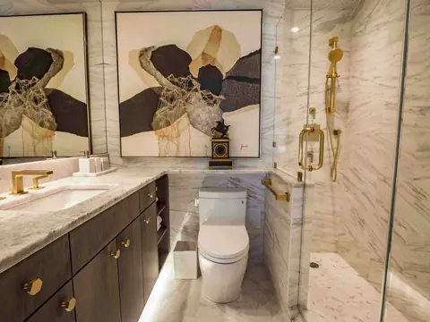 A marble bathroom with a dark vanity on brass knobs, a brass faucet, two abstract prints and a glass shower with brass fittings