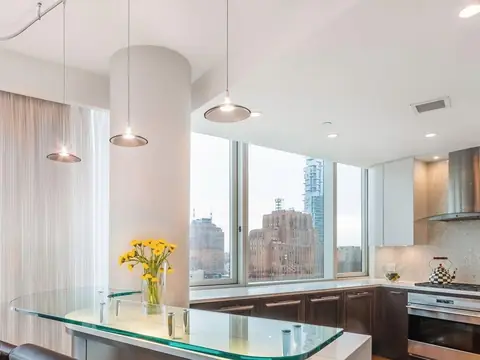 A high-rise kitchen with a glass breakfast counter, pendant lights, a round column and windows onto city towers