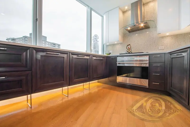 A long run of dark base cabinets with a built-in oven, amber lighting under the toe kick and windows above the counter