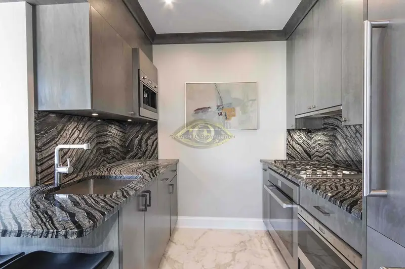 A galley kitchen with grey flat cabinets, black and white wave-patterned stone counters and backsplash and a marble floor