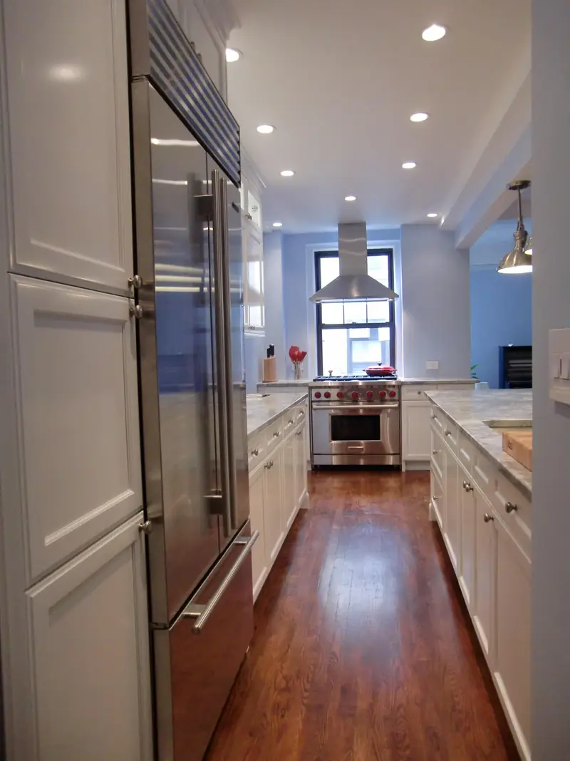 A galley kitchen with white cabinets, a steel refrigerator, a range under a hood at the far window and a cherry floor