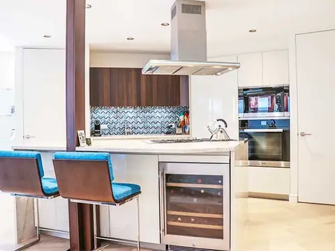 A kitchen breakfast bar with two blue-cushioned stools, a wine refrigerator, a walnut column and a chevron tile backsplash