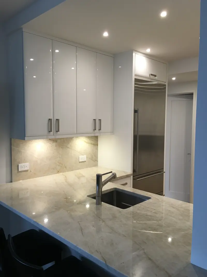 A kitchen corner with a polished stone counter and backsplash, lit white lacquer wall cabinets and a steel refrigerator