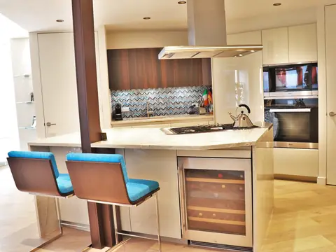A kitchen with cream cabinets, walnut upper cabinets, a chevron tile backsplash, an island hood and two blue stools at the breakfast bar