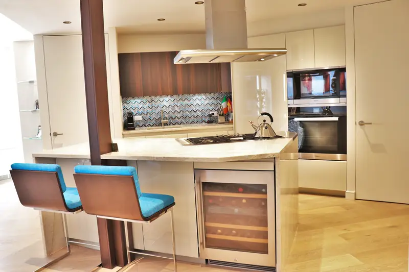 A kitchen with cream cabinets, walnut upper cabinets, a chevron tile backsplash, an island hood and two blue stools at the breakfast bar