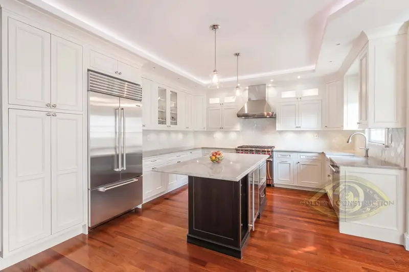A kitchen with white cabinets to the ceiling, a dark island with a white top, a steel refrigerator, two pendants and a cherry floor