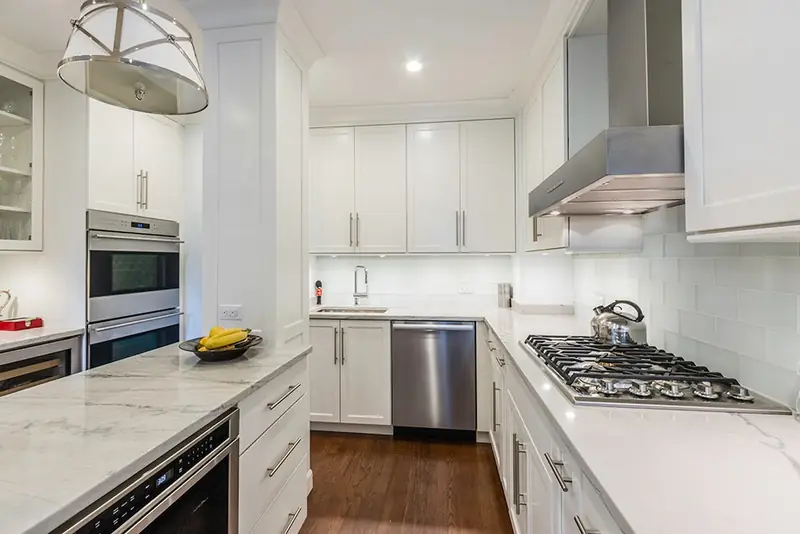 A white kitchen with marble counters, a gas cooktop under a steel hood, a double wall oven and a drum pendant light