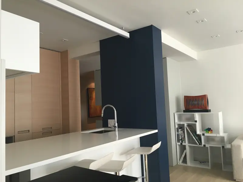 A kitchen with a long white counter and sink, pale wood cabinets, a navy blue column and white stools