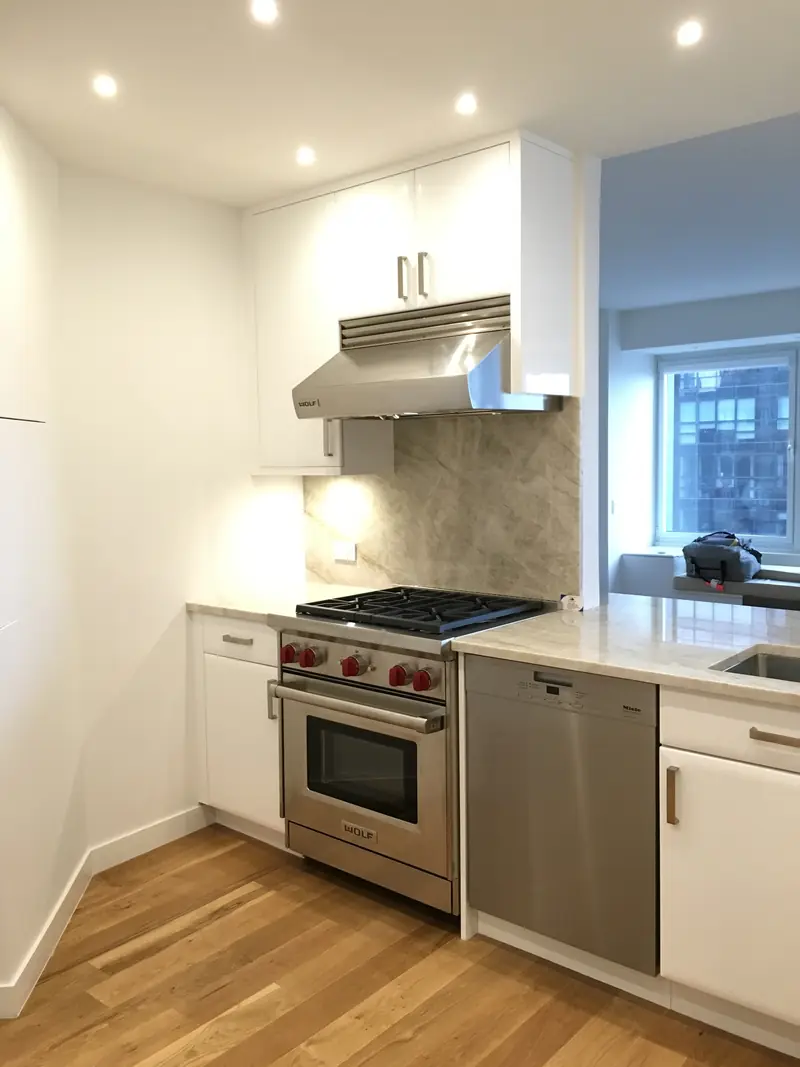 A kitchen with white lacquer cabinets, a professional range with red knobs under a steel hood and a stone backsplash