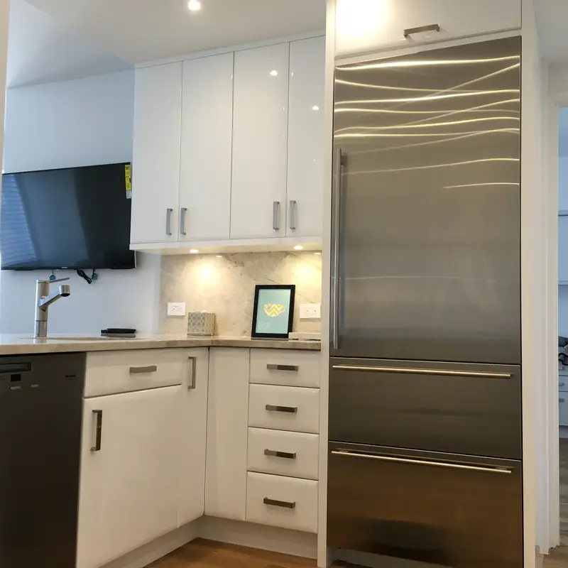 A kitchen corner with white lacquer cabinets, a stone counter and a steel refrigerator with a brushed wave pattern