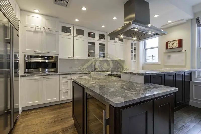 A kitchen with white shaker cabinets and glass-front uppers, a dark island under a grey granite top and a steel island hood