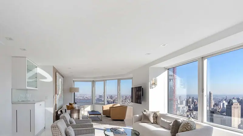 A white living room with windows onto a skyline, a sofa and armchairs around a glass table and white built-in cabinets