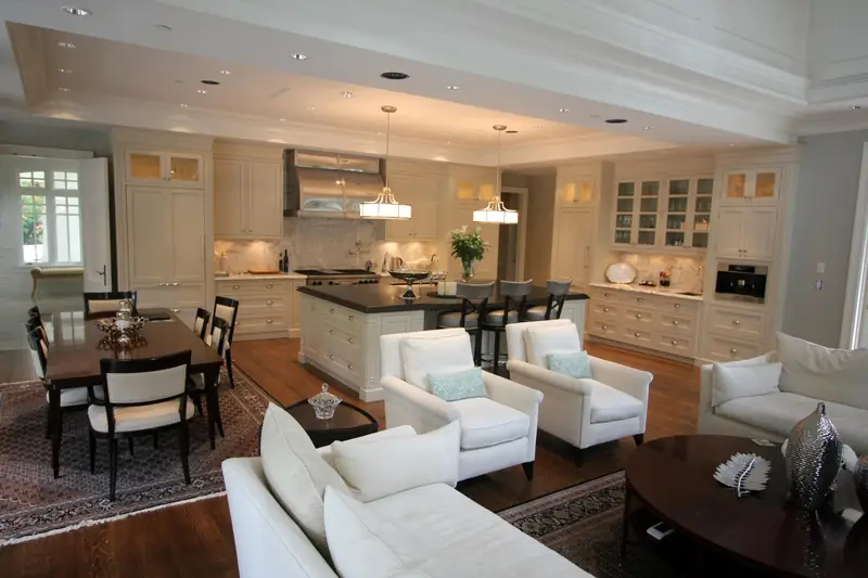An open living, dining and kitchen room with cream cabinets to the ceiling, a dark island with stools and two pendant lights